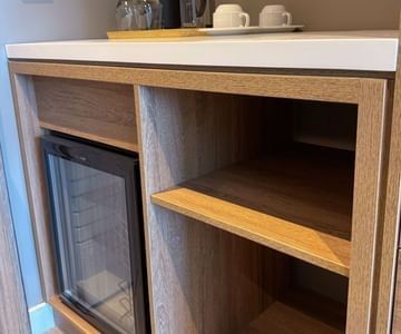 Mini fridge inside a wooden cabinet with coffee cups and glassware on the counter in a guest room at modern Unika Hotels