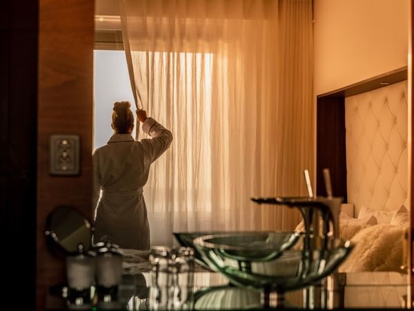 Person in bathrobe opening sheer curtains in warm-toned hotel room with soft morning light streaming through window