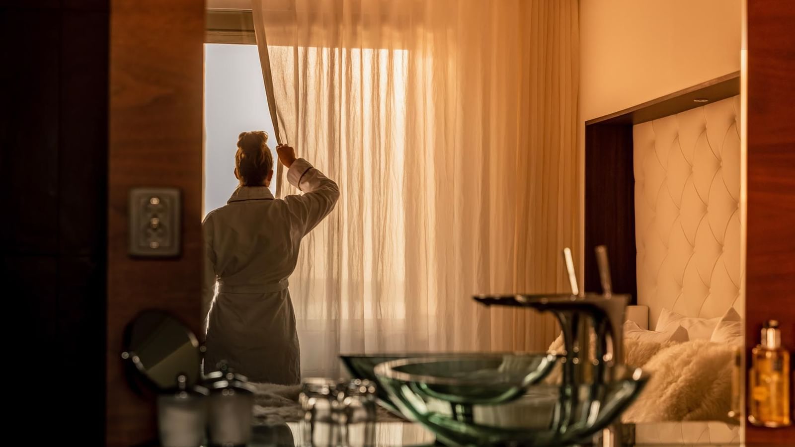 Person im Bademantel öffnet morgens die Vorhänge in einem warm beleuchteten Hotelzimmer mit elegantem Interieur