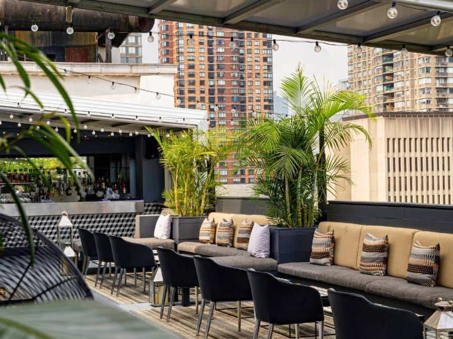 A rooftop bar with a long sofa, chairs, and a bar counter, surrounded by plants and city views.