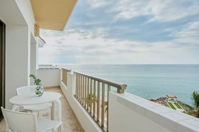 Balcony with white table and chairs, overlooking ocean and beach with palm trees.