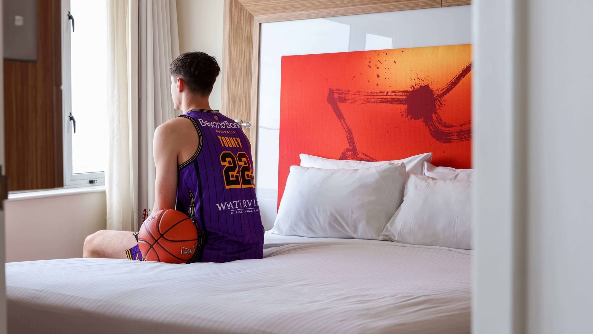 Basketball player sitting on the bed and gazing out the window in Superior Spa Suites at Novotel Sydney Olympic Park