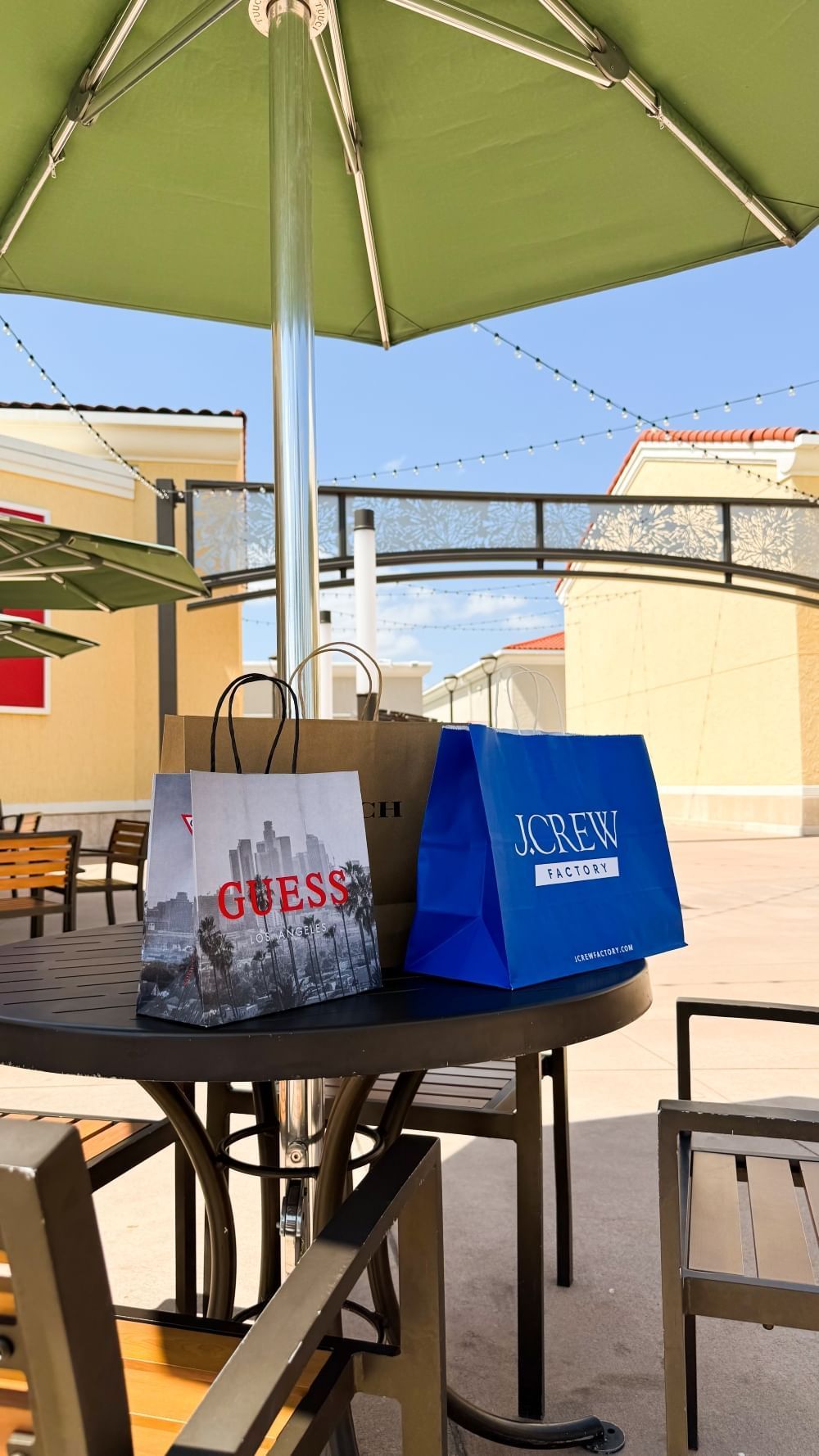 A table with three shopping bags underneath an umbrella with buildings of an Orlando outlet mall in the background.