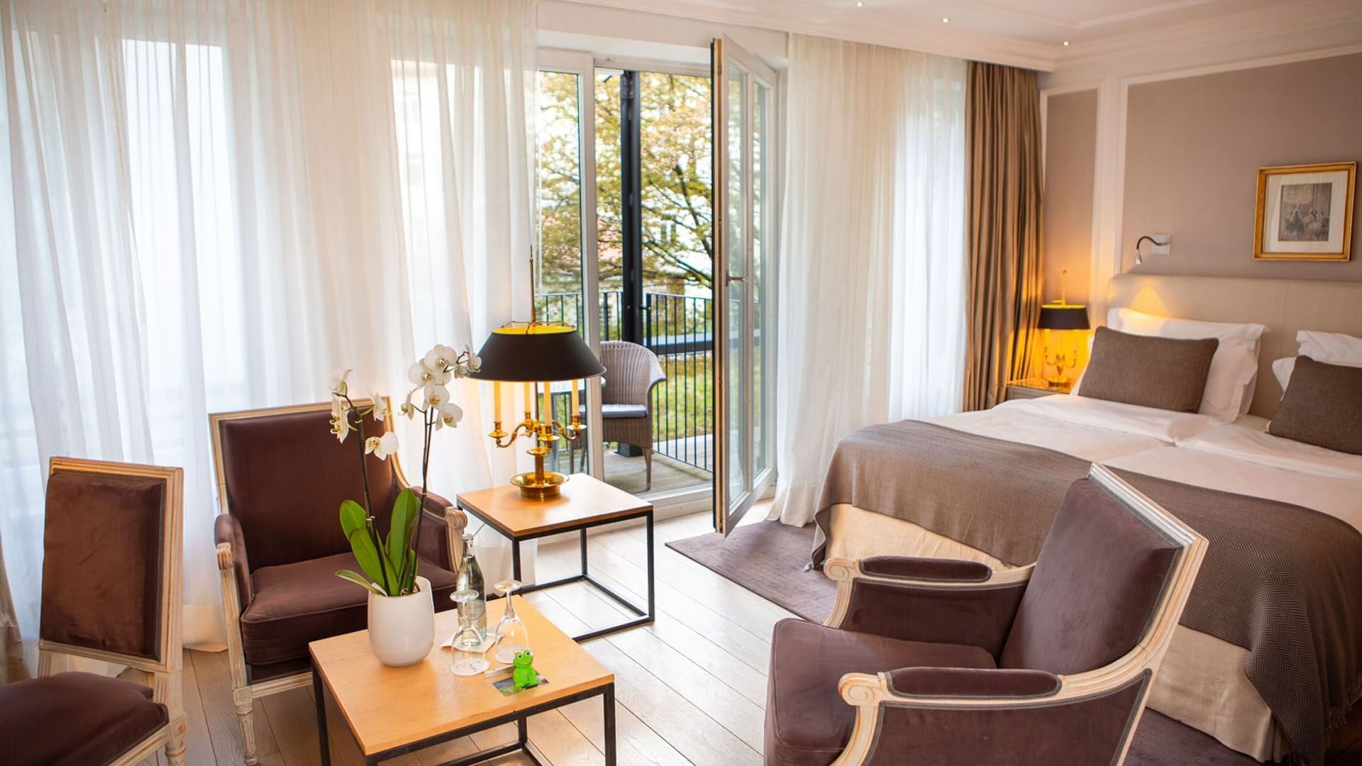 Bed with brown armchairs and wooden tables, by the balcony door in Deluxe Garden Double Room at Hotel Palace Munich
