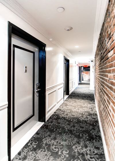 East Village Hotel hallway with carpeted floor, white walls, black doors, and a brick wall on the right.