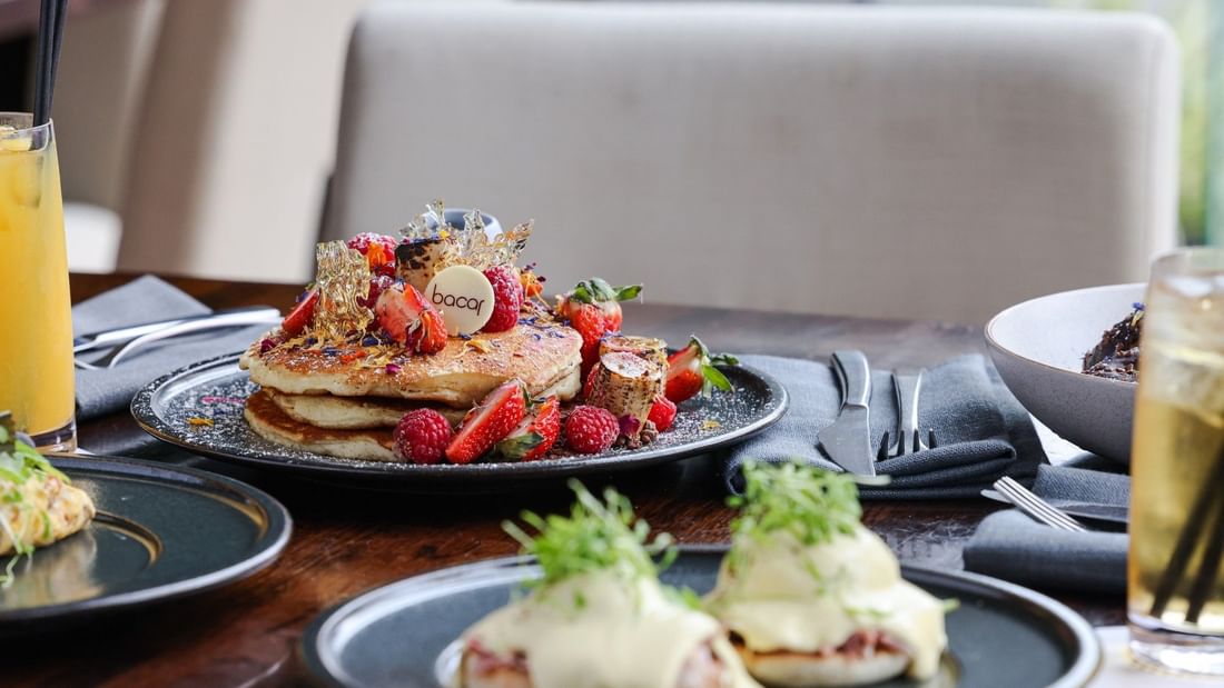 Gourmet breakfast spread featuring pancakes topped with fresh berries and eggs benedict on dark plates with beverages