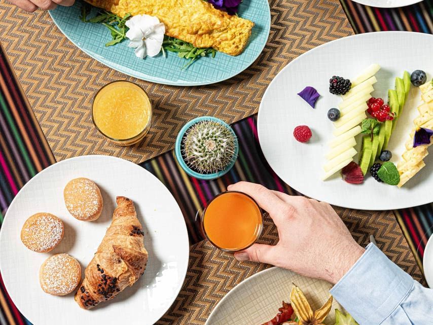 Brunch spread featuring an omelet, croissant, pastries, fresh fruit, and drinks on a patterned tablecloth at The Glam Hotel