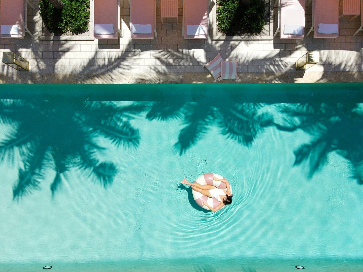 Aerial view of a pool with a woman floating on a pink and white pool float, surrounded by pink lounge chairs and palm trees.