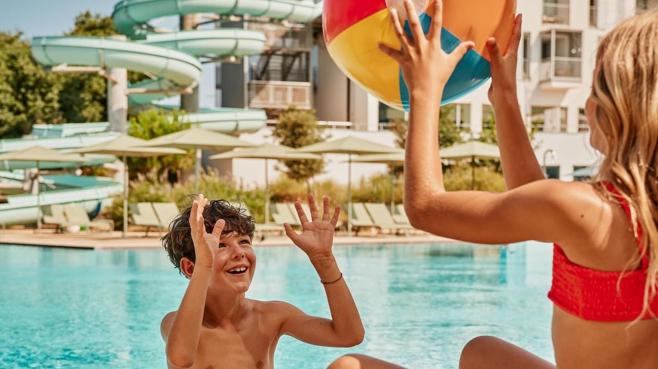Bambino e bambina sorridenti giocano con una palla sul bordo della piscina, con scivoli e ombrelloni sullo sfondo.