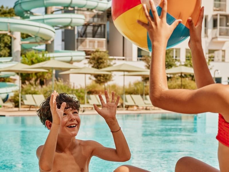 Zwei Kinder spielen mit einem bunten Beachball am Poolrand neben einer Wasserrutsche.