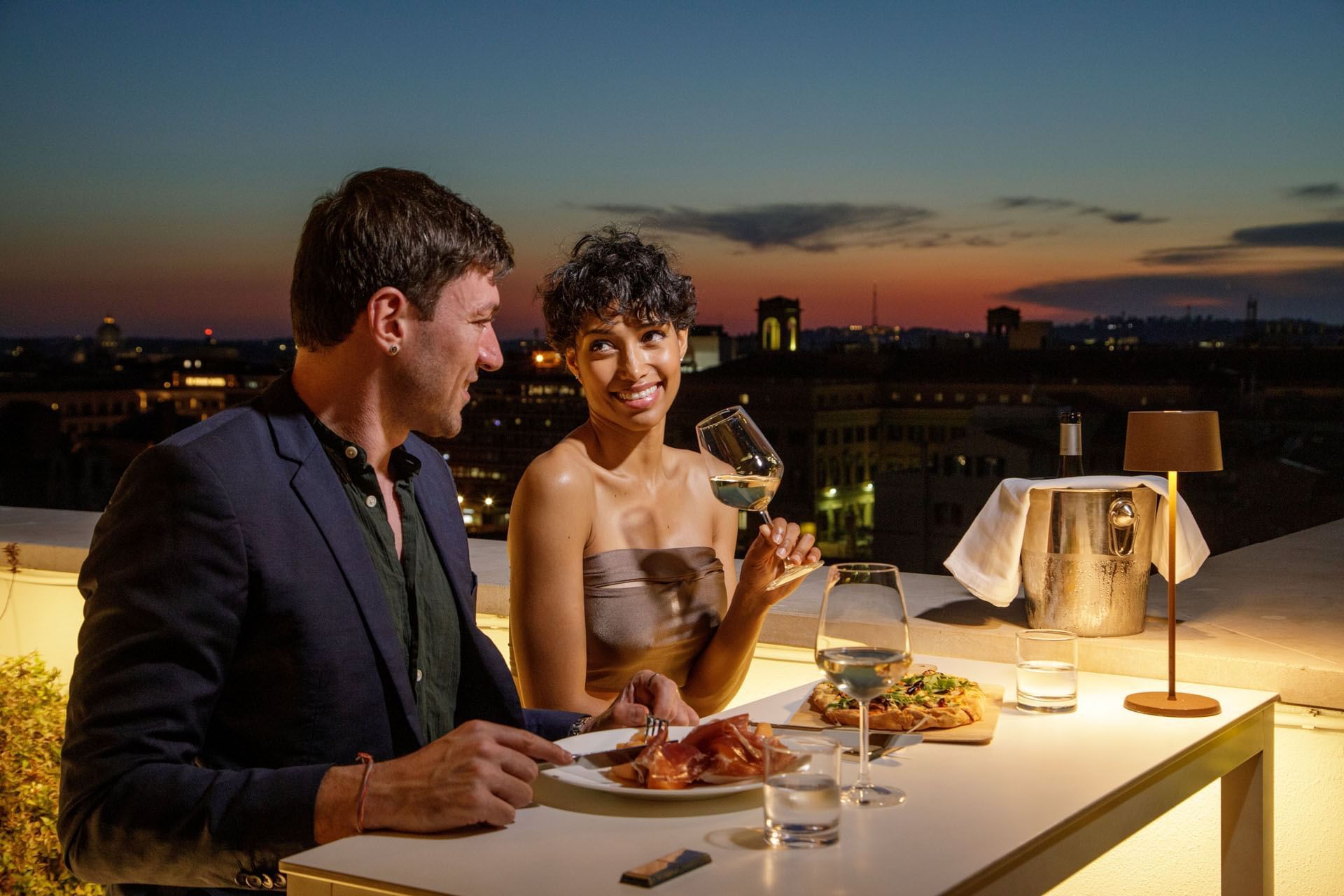 Couple enjoying a romantic rooftop dinner with wine and a sunset city view at The Independent
