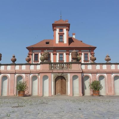 Pink and white historic building with a central brown door and decorative vases on the roof.