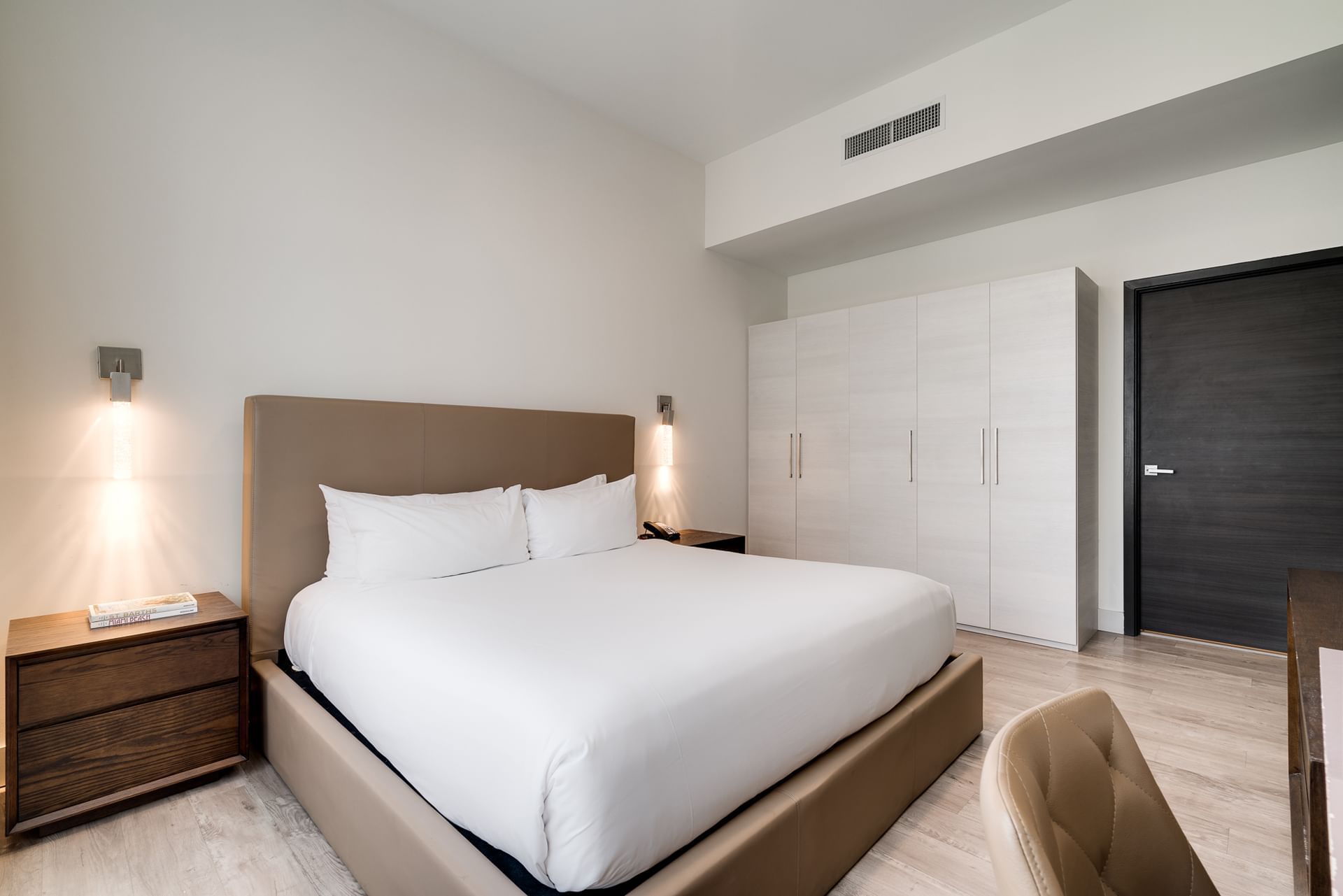 A modern hotel room with a large bed, nightstand, wardrobe, and chair.