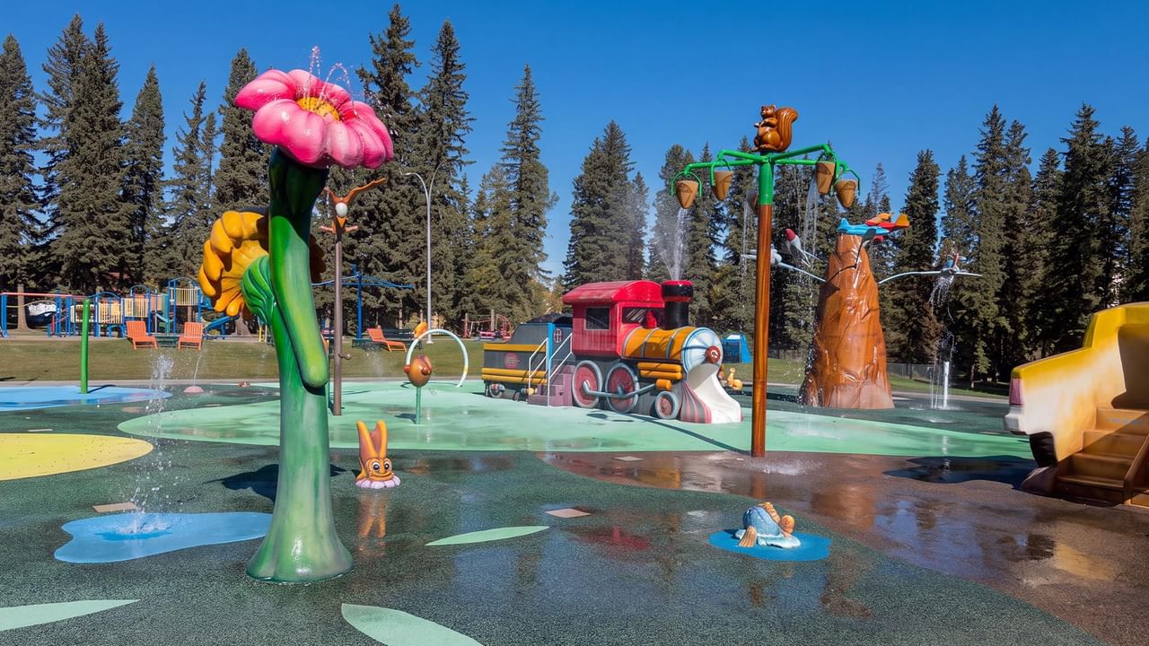 Kinsmen Spray Park features colorful water play structures including a pink flower, train, and tree surrounded by evergreens.