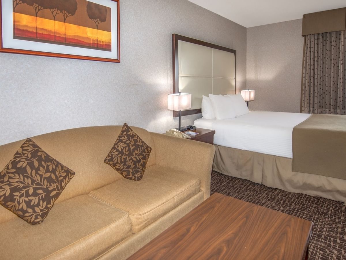 Hotel room with couch, coffee table, bed, window, and wall art of sunset over trees.