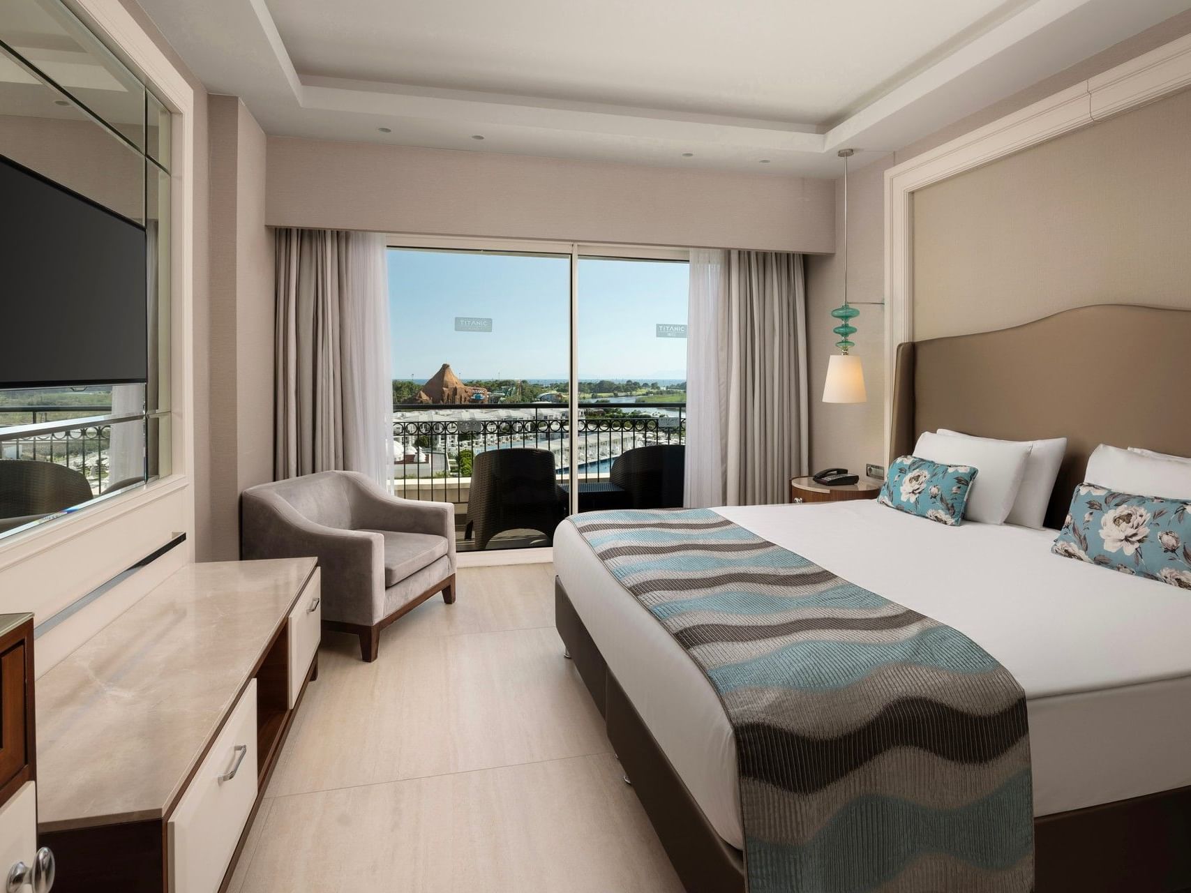 Modern hotel room with king bed, balcony view, wall-mounted TV, and neutral decor at Titanic Deluxe Golf Belek