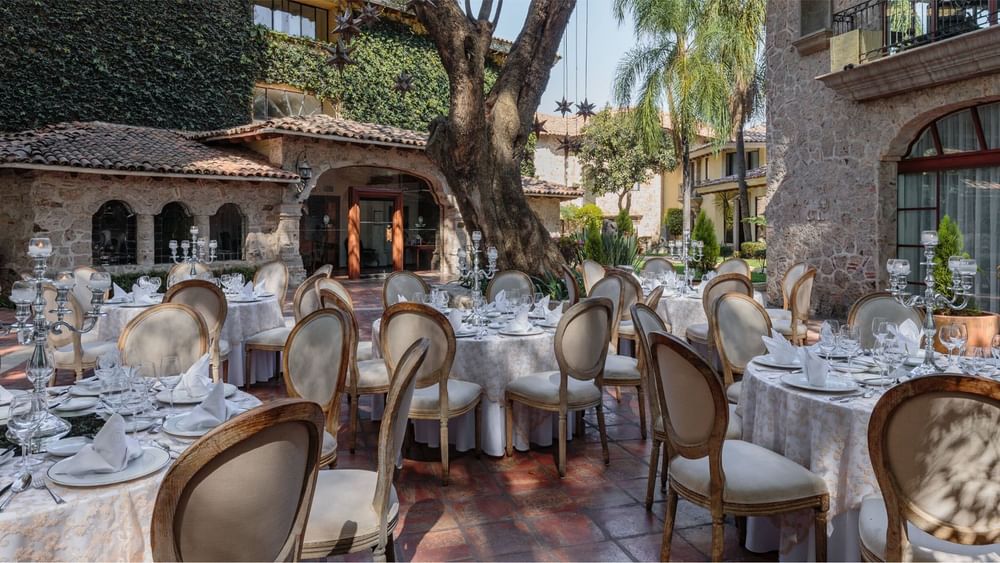 Salón Virreinal outdoor dining setup with round tables and white linens under large trees at Quinta Real Guadalajara