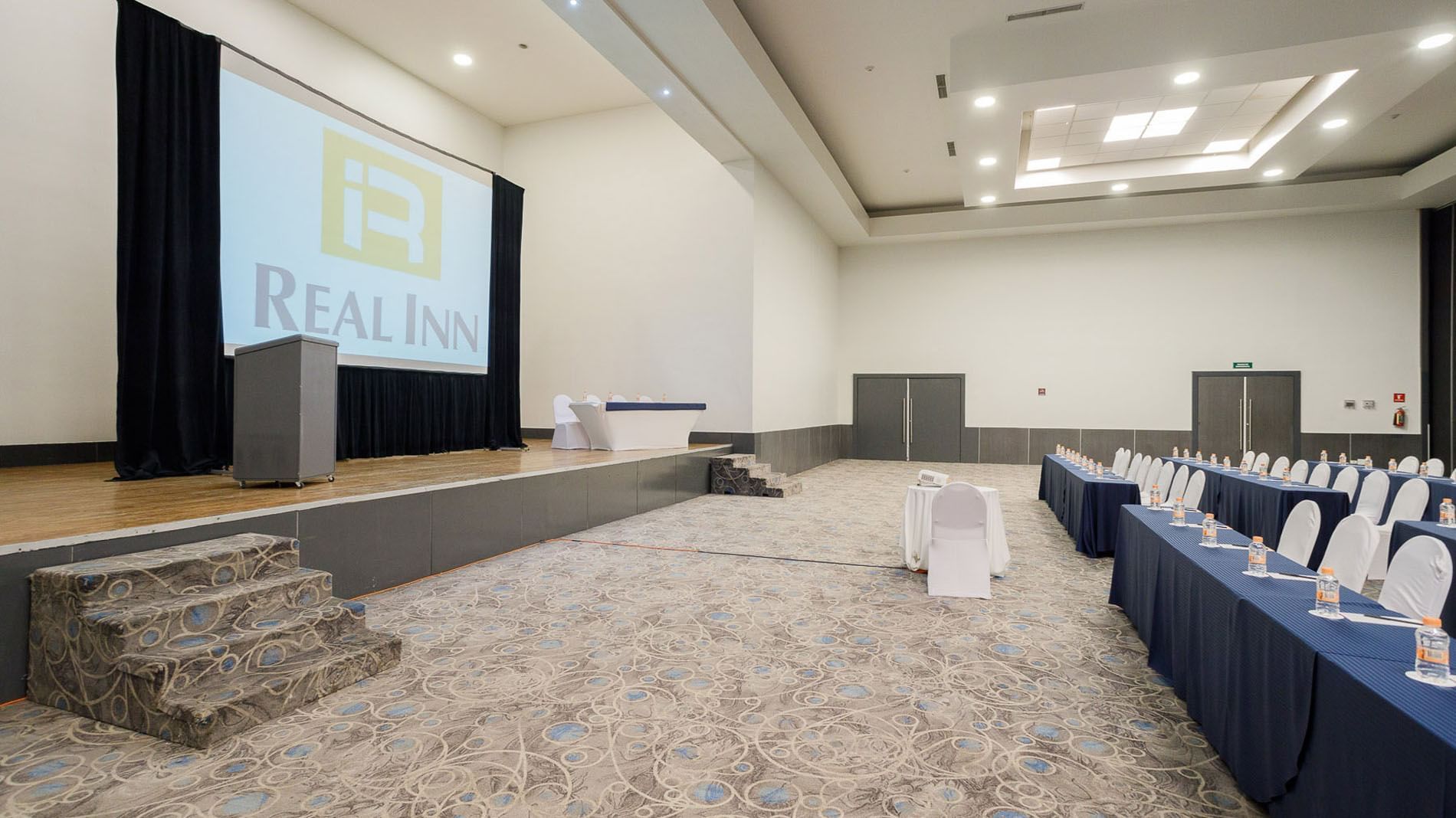 Classroom set up facing the projector screen with a stage in the Nápoles meeting hall at Real Inn Tijuana