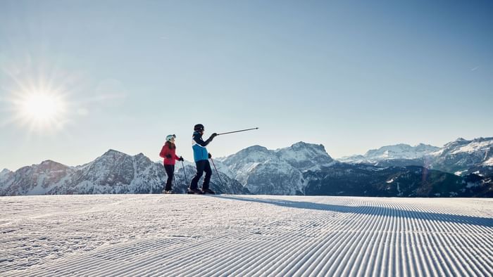 Due sciatori su neve battuta con montagne innevate sullo sfondo e sole splendente.