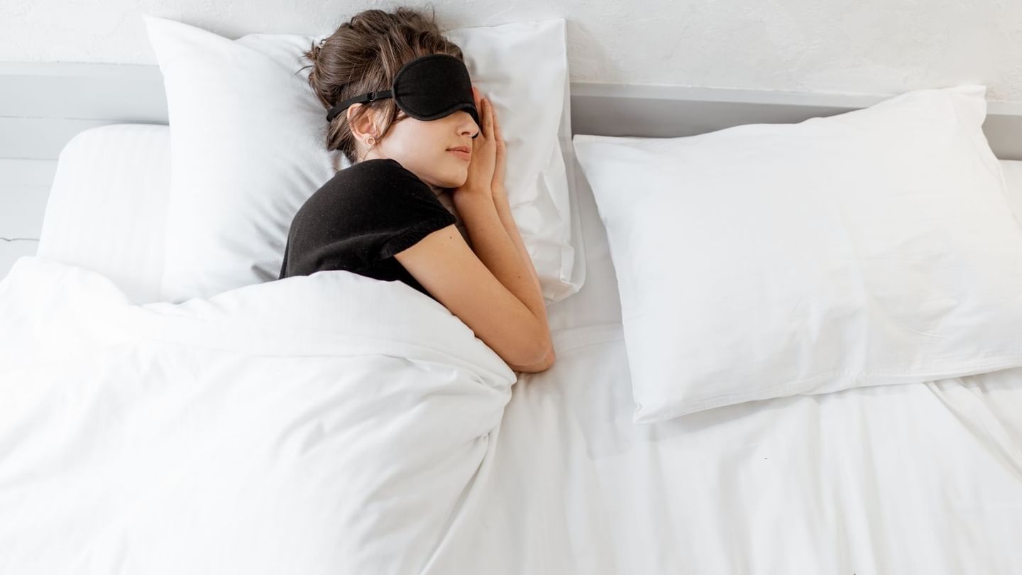 Woman lying in bed with white bedding and pillow, wearing a black eye mask, resting her head on a pillow.