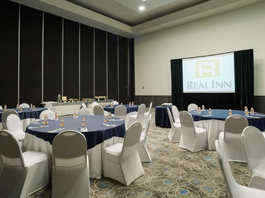 Milán Verona meeting room with banquet set-up, white chairs, and a projector screen at Real Inn Tijuana