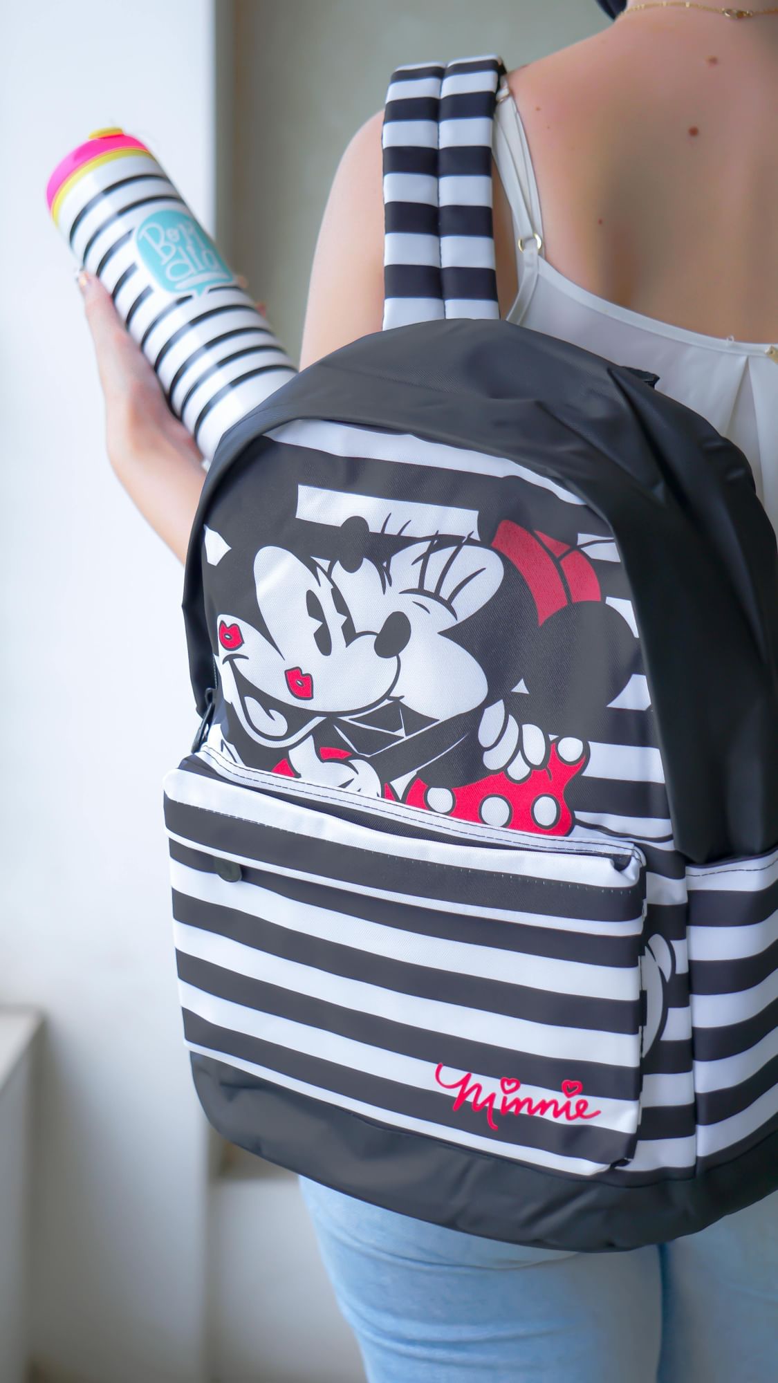A woman in a white tank top carries a black and white striped backpack on her back with an image of Minnie Mouse kissing Mickey Mouse.