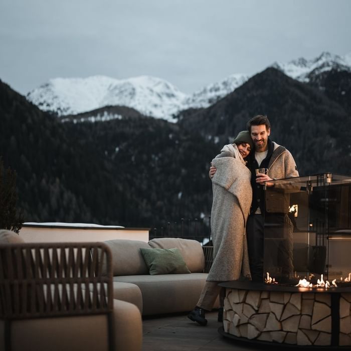 Coppia avvolta in coperte vicino a un fuoco esterno con vista sulle montagne innevate presso Falkensteiner Hotel Falkensteinerhof