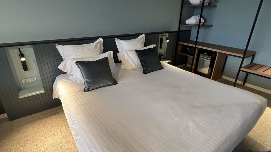 Chambre d'hôtel avec grand lit blanc, oreillers, et mobilier moderne dans un décor bleu et gris.