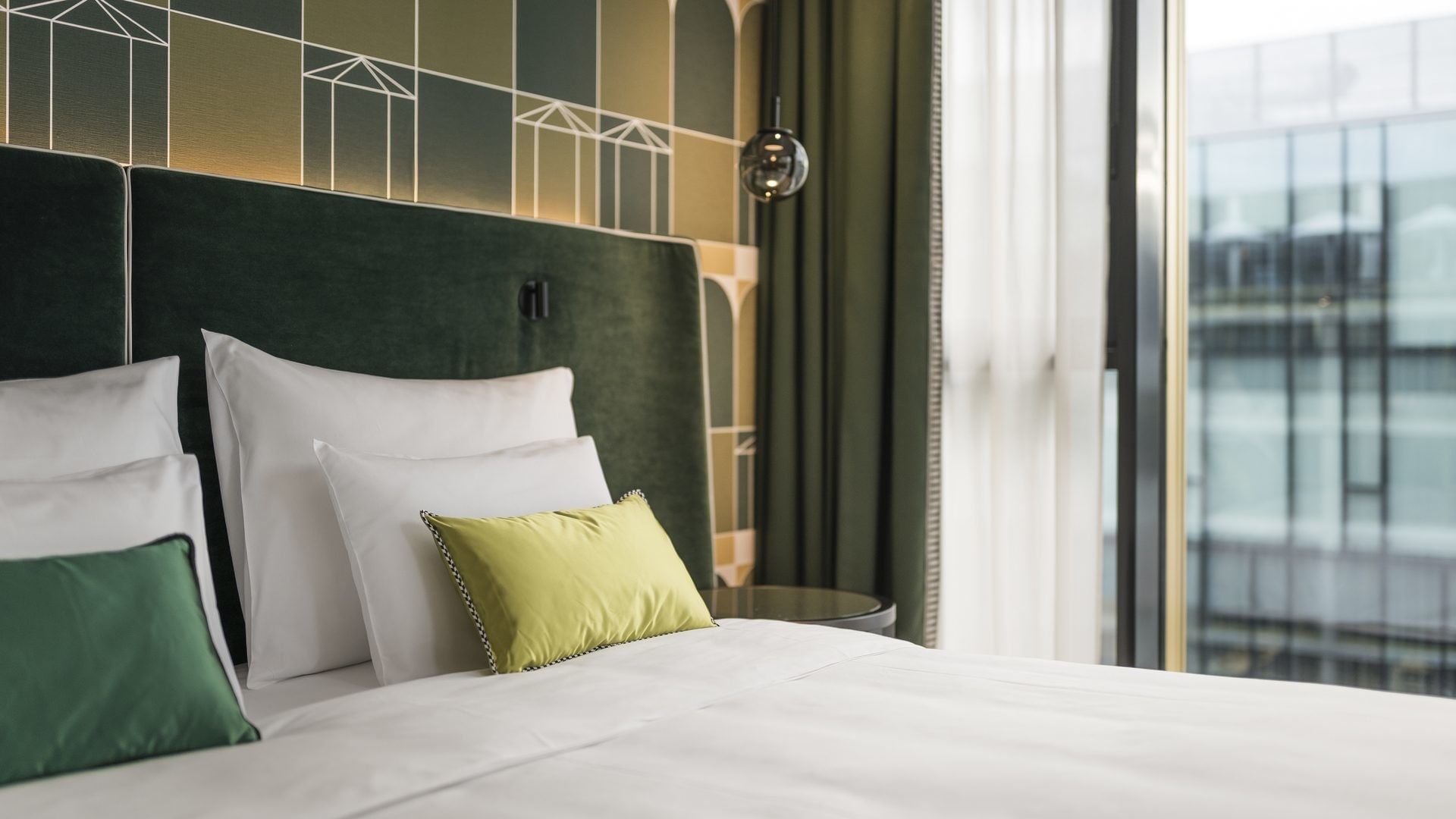 A hotel room with a white bed, three white pillows, one green pillow, and a patterned wall.