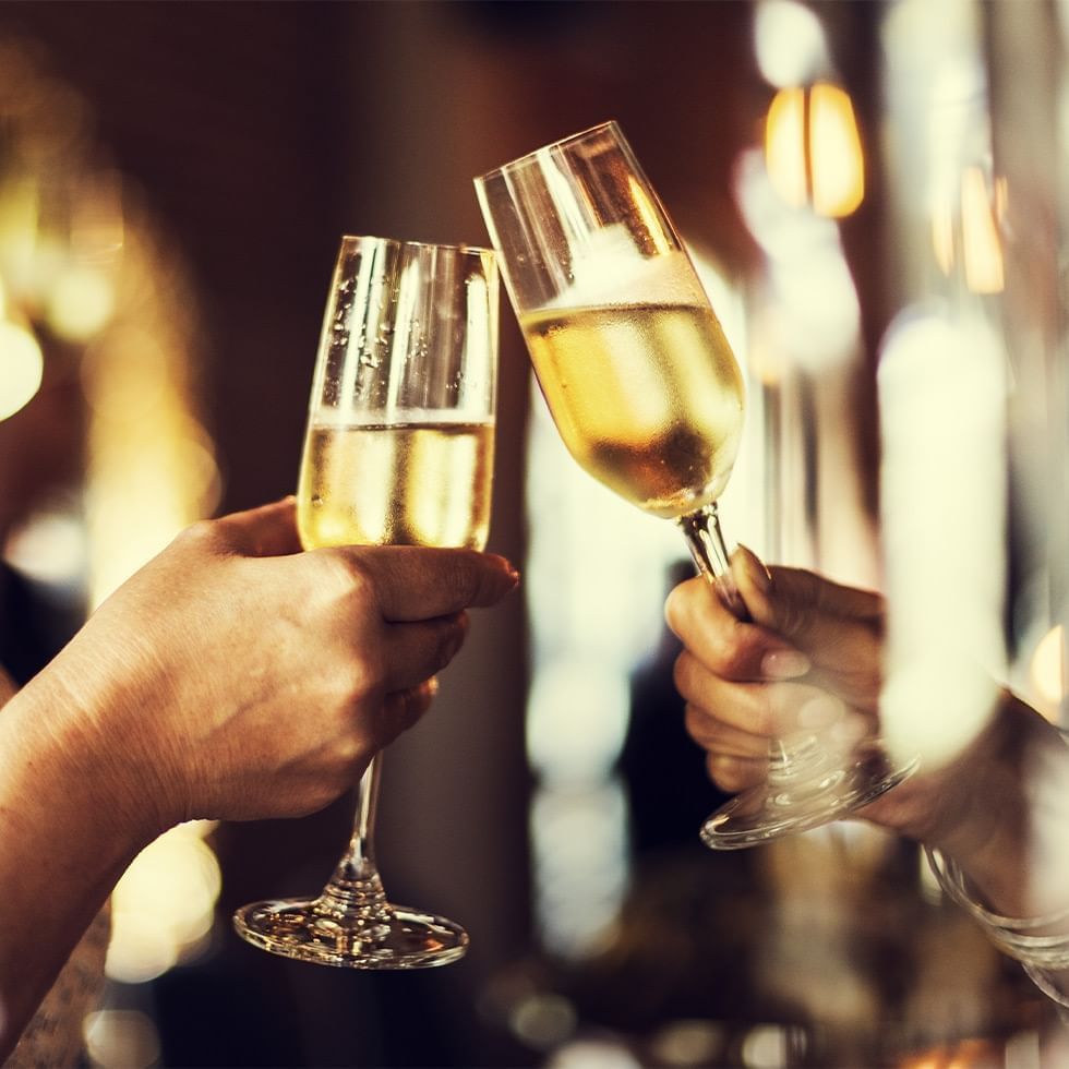 Due mani sollevano calici di champagne per un brindisi, con sfondo sfocato di luci calde.