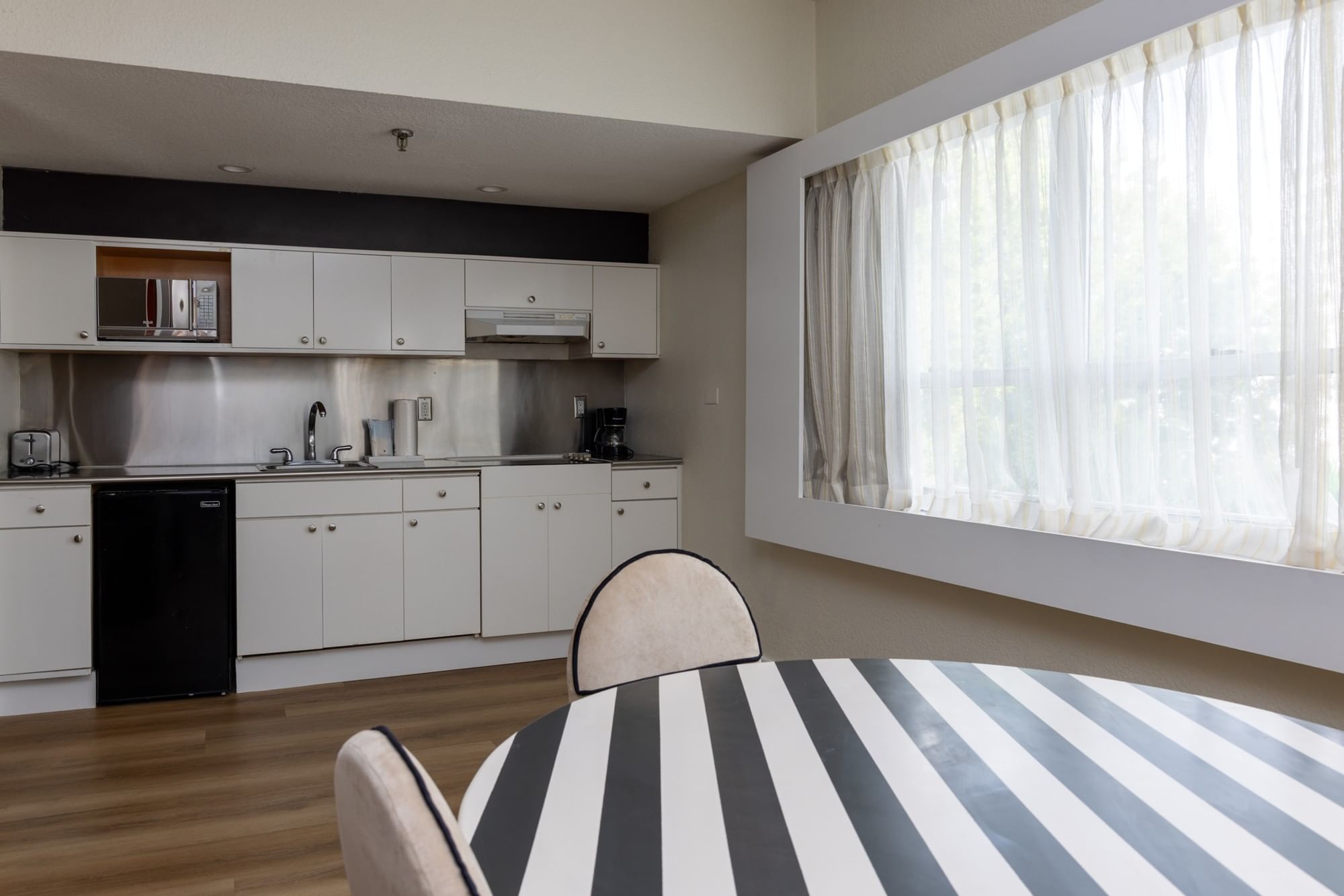 A kitchenette with white cabinets and black appliances, a dining area with a striped tablecloth and two chairs, and a large window with sheer curtains.