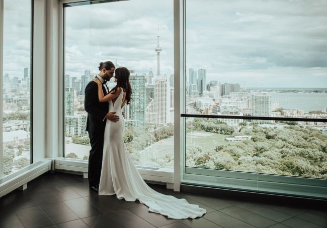 Bride and groom embracing beside floor to ceiling glass facing Toronto skyline with CN Tower landmark at Hotel X Toronto