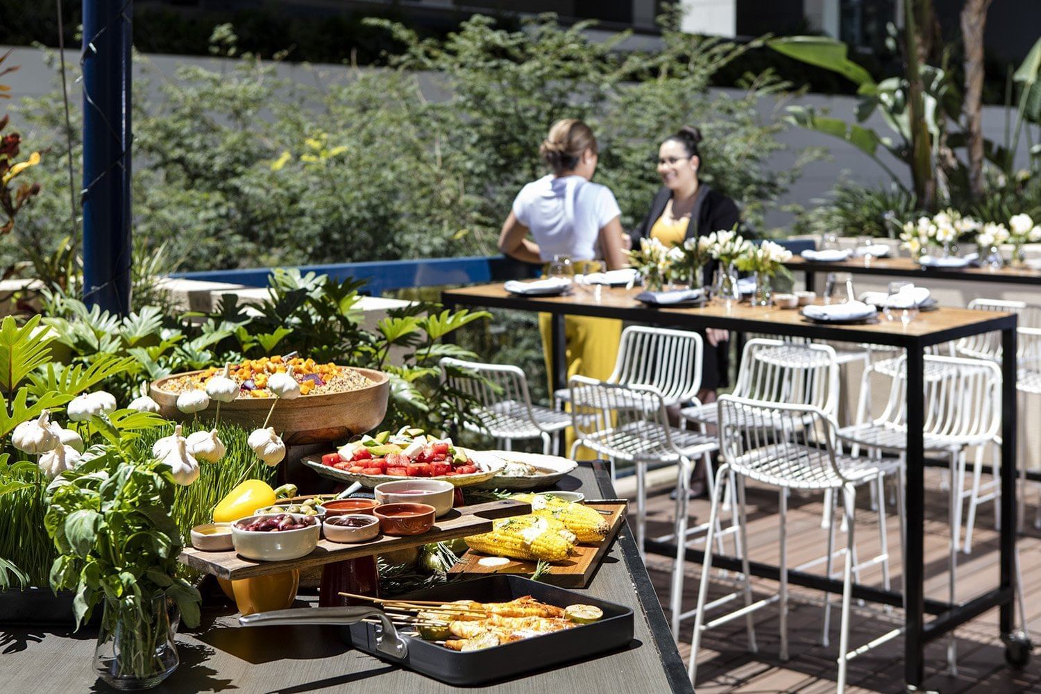 Outdoor buffet with a grilling station, fresh vegetables, and two guests chatting at a high table inside The Sebel Brisbane
