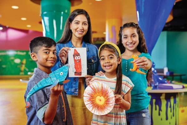 A woman and three children stand smiling and presenting handmade artwork in front of a colorful backdrop. 