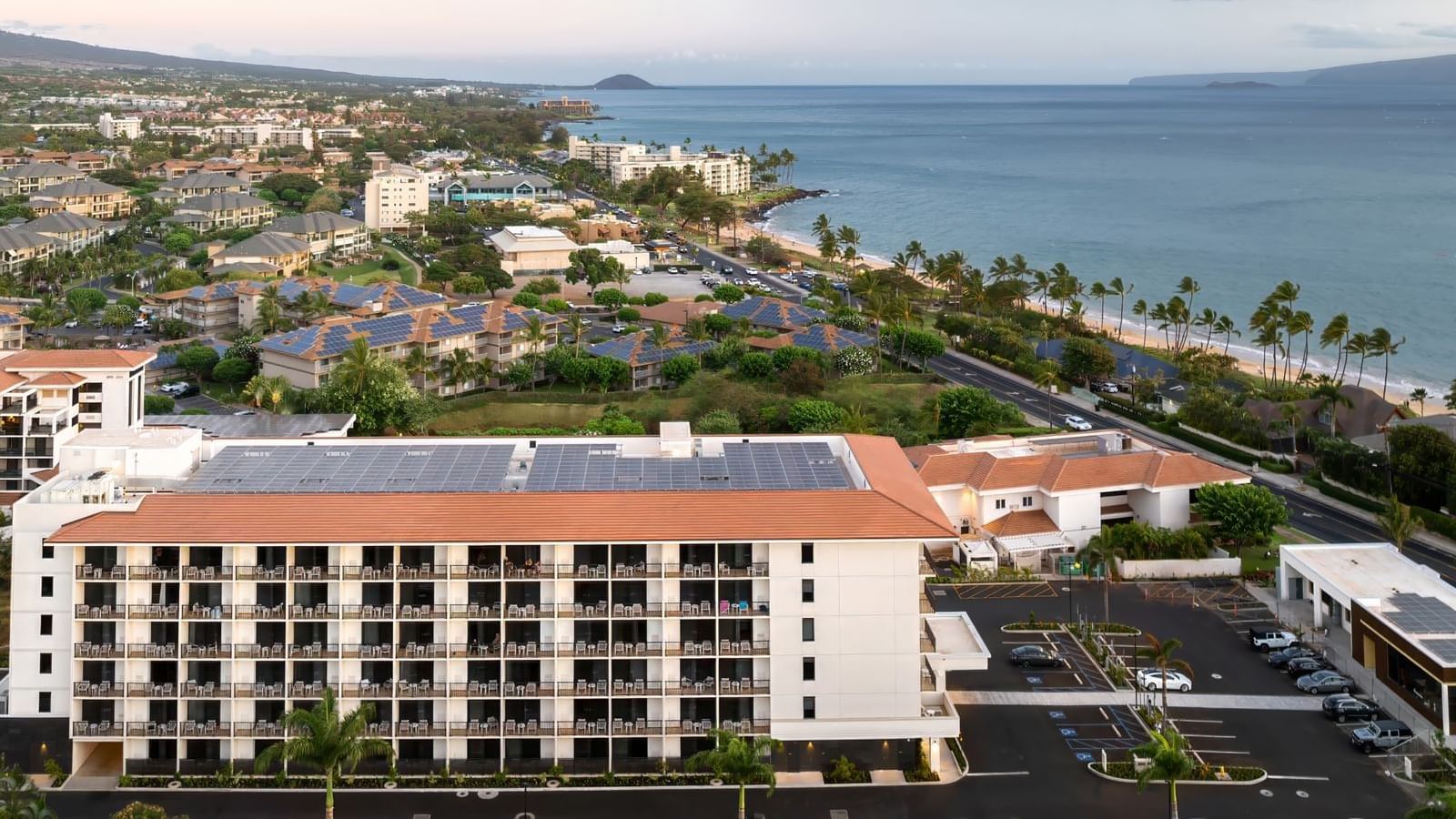 An aerial view of a large white building with an orange roof and solar panels, surrounded by a parking lot and ocean views.