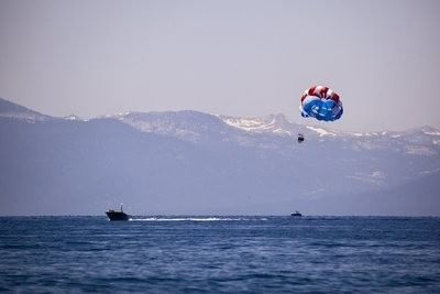 A blue parachute carries a parasailer high above Lake Tahoe with snow-capped mountains behind near Americana Village Suites