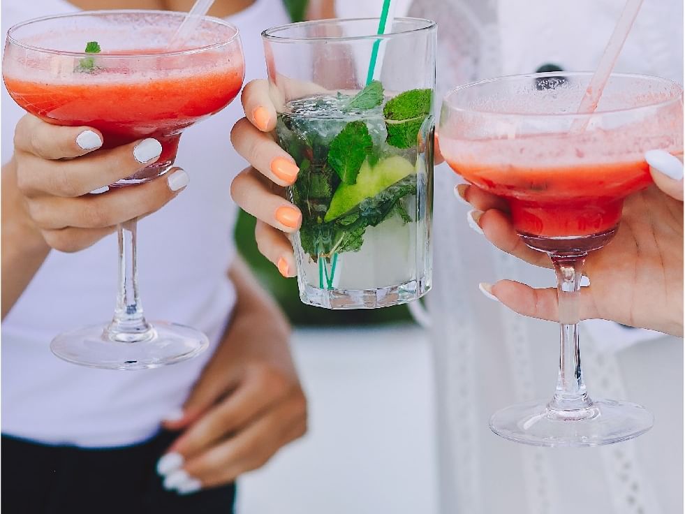 Three people holding martini glasses with drinks and straws, two with red drinks, one with green.