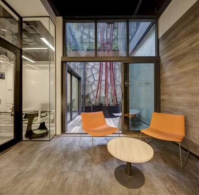 Lounge area with two orange chairs, a round table, and large windows overlooking a green courtyard in Room D at Hotel Nodo