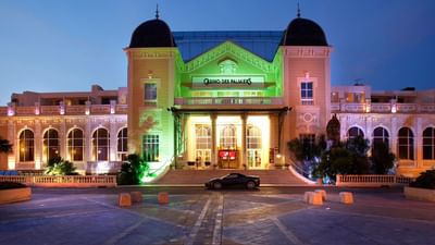 Nighttime view of Casino des Palmiers with green and yellow lights, black car parked in front.