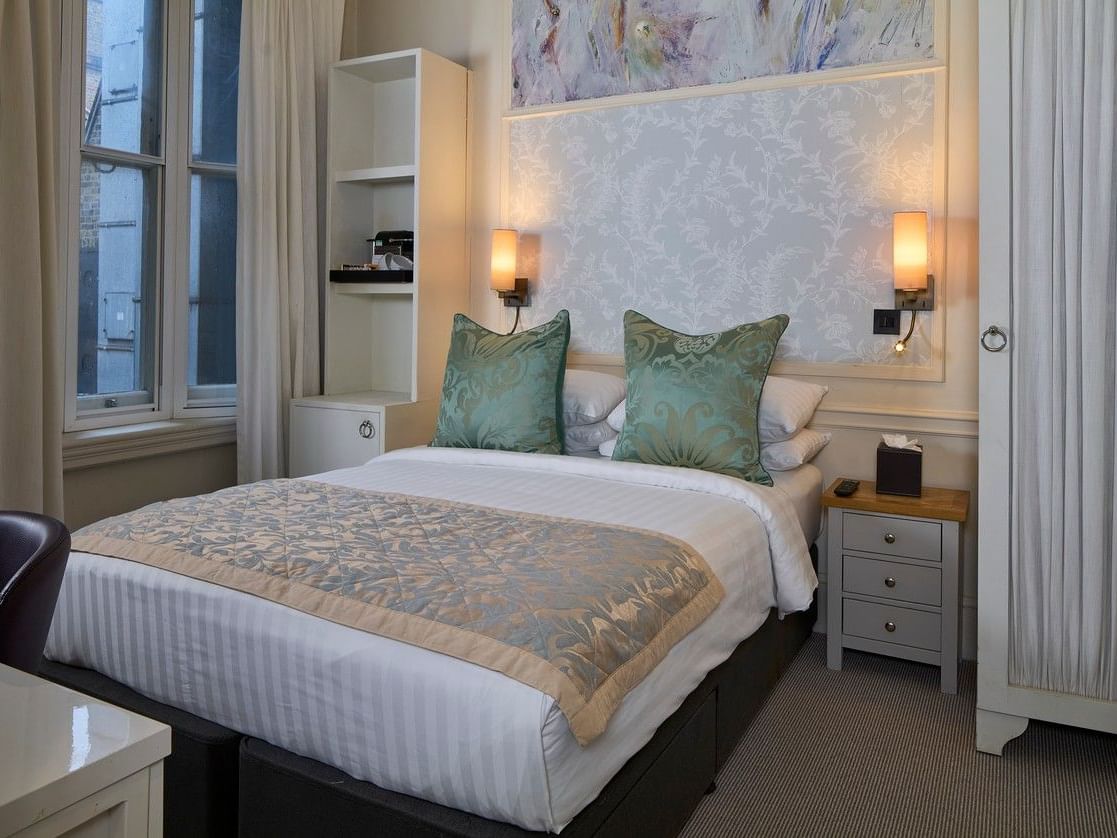 Small double room with upholstered bed, decorative cushions, bedside lamps, and window at Sloane Square Hotel.