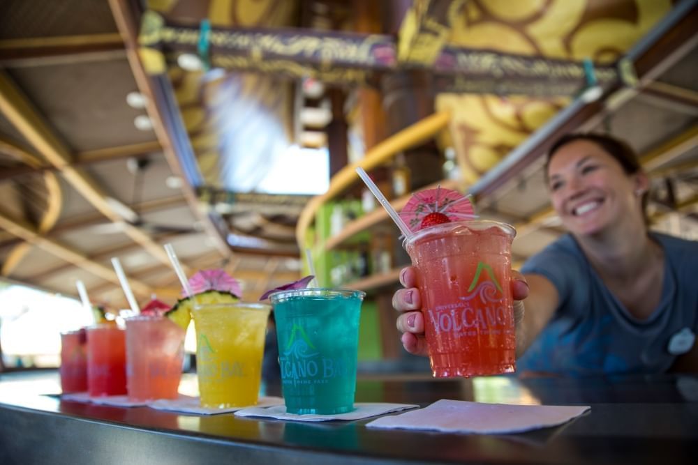 A waitress places a pink cocktail in a plastic Volcano Bay cup on the end of a row of colorful cocktails. 