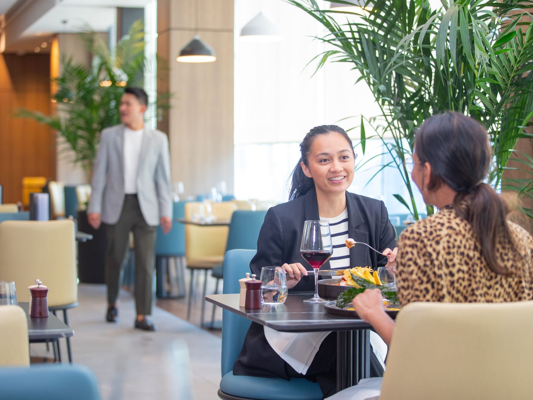 Two women having a meal at a restaurant with a man walking past in the background.
