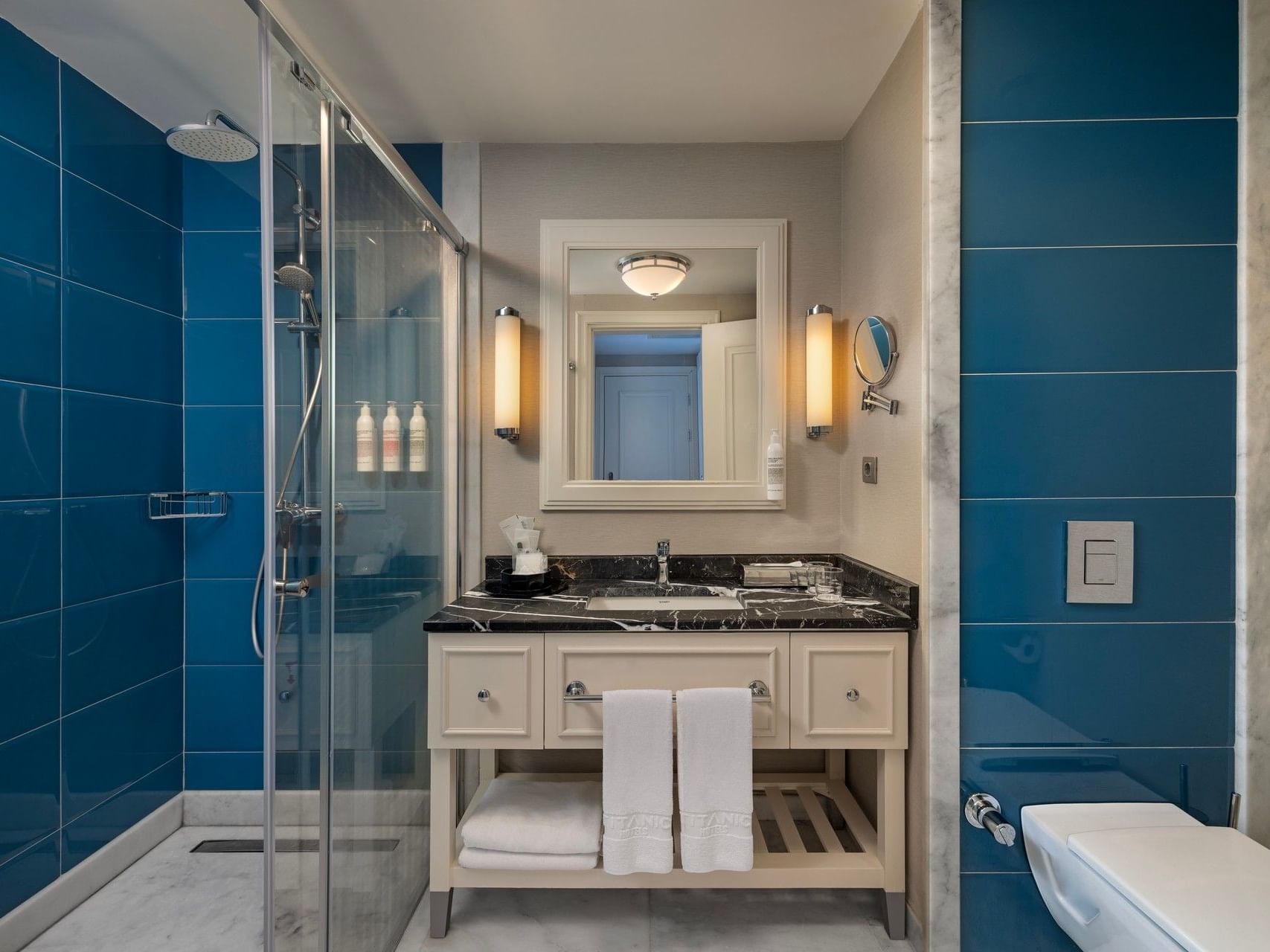 Modern bathroom with blue tile walls, glass shower, white vanity with dark countertop, and illuminated mirror at Titanic Deluxe Golf Belek