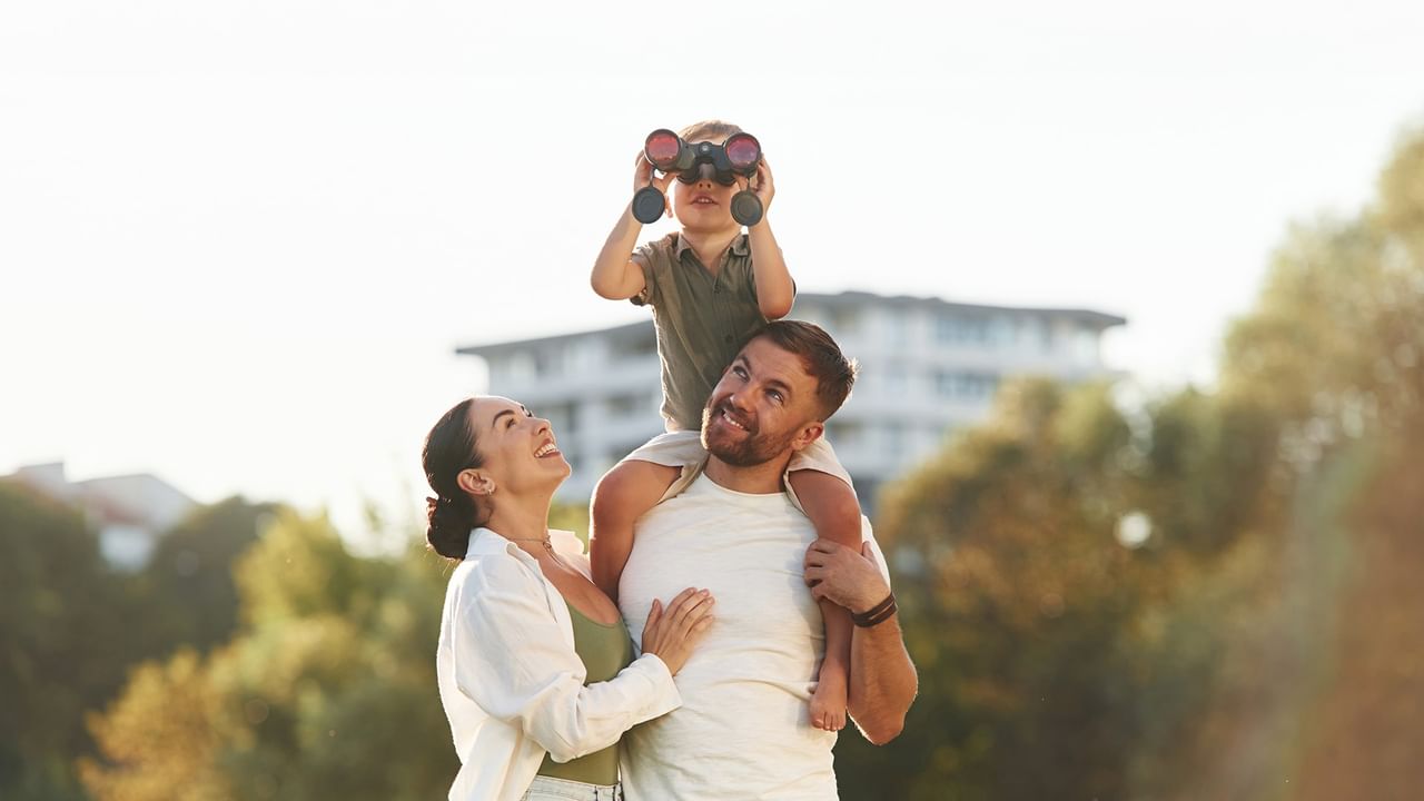 A man and woman hold a child with binoculars on their shoulders in an outdoor setting.
