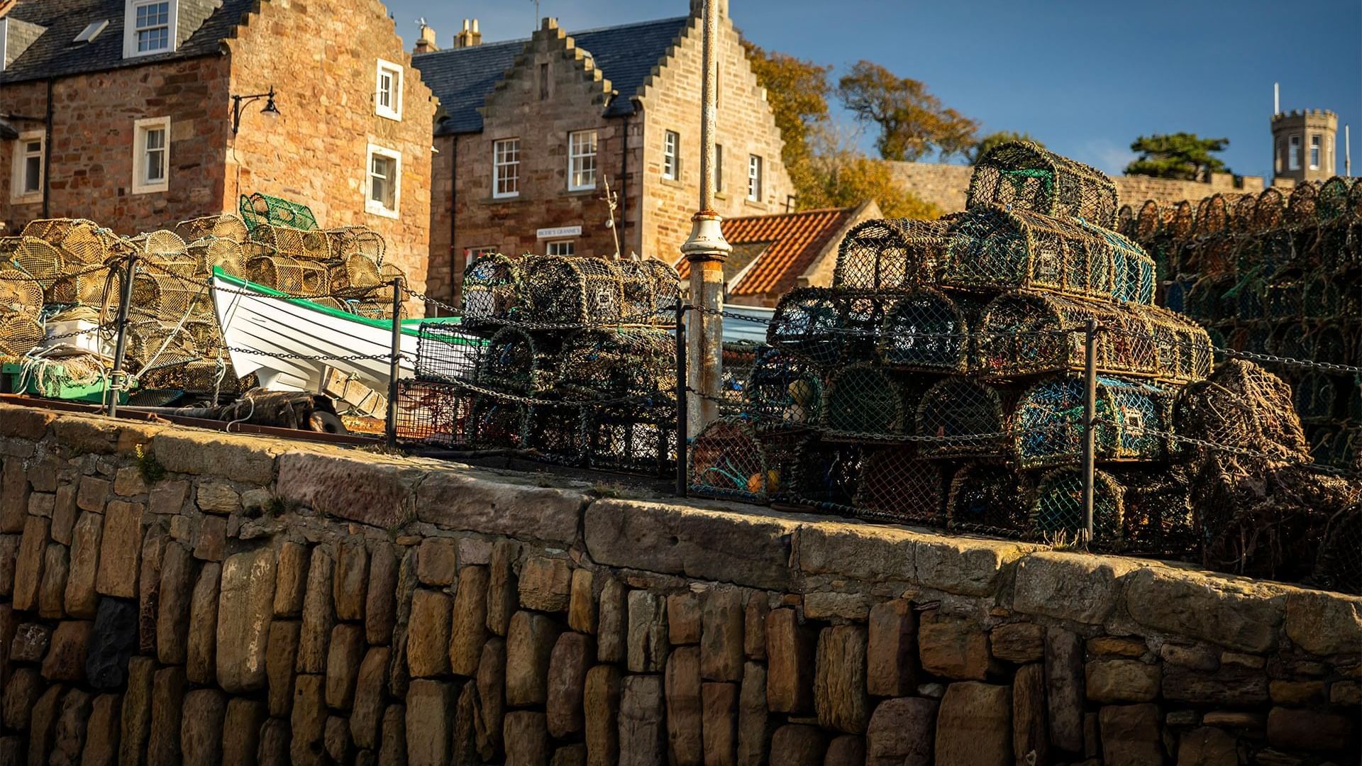 Fishing nets and traps stacked by a stone wall near traditional cottages and a boat at Seaton House