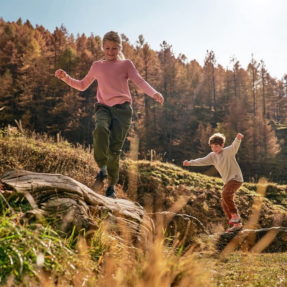 Bambina e bambino saltano su rocce in un bosco autunnale.