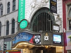 Opera House marquee with Les Miserables poster and lit lights, flanked by other buildings and street signs.