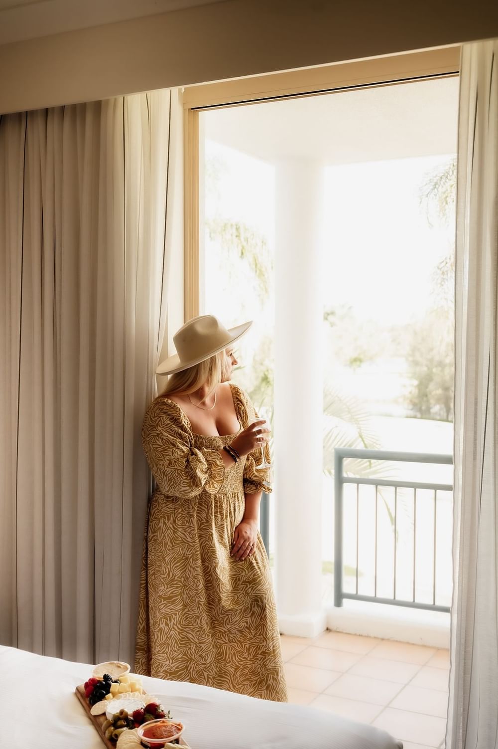 Woman in hat and dress standing by balcony door with charcuterie board on a bed.