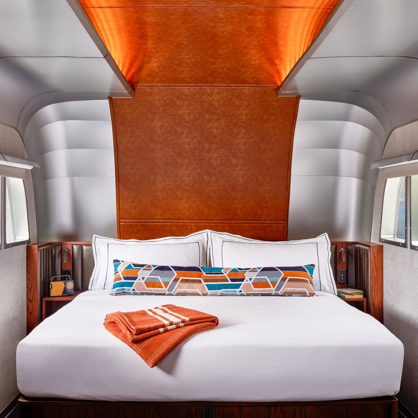 Retro Airstream trailer bedroom with white bedding, geometric orange pillows, and curved metallic walls with windows.