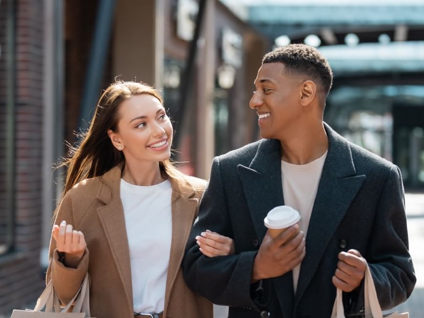Smiling man and woman walking, holding coffee and shopping bags, in an urban setting.
